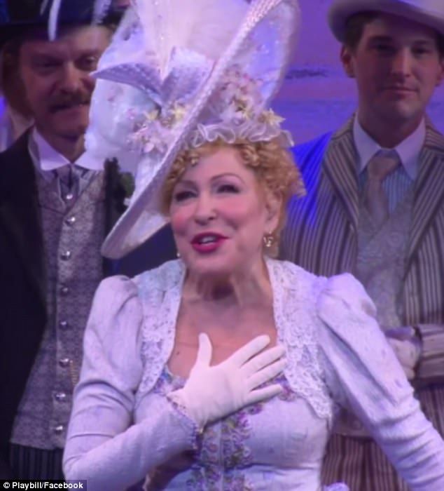 Bette Midler in a Victorian-style lavender dress with white gloves, a decorative hat, and lace, touching her chest on stage.