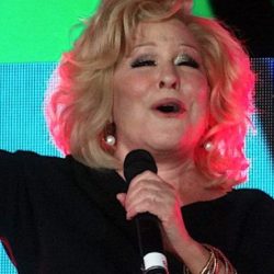 Older woman singing on stage into a microphone, wearing a black dress and gold bracelets.