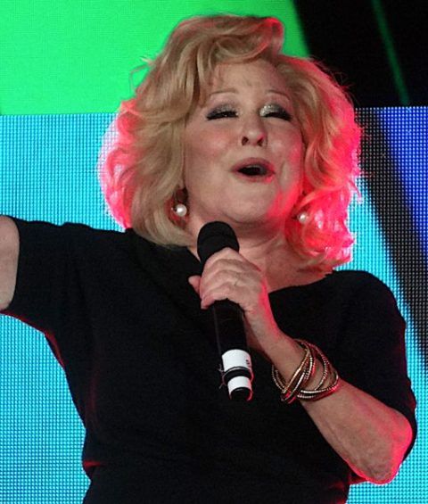 Older woman singing on stage into a microphone, wearing a black dress and gold bracelets.