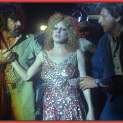 Woman in a glittery, sparkling gown is being escorted by two men who hold her arms in a dim backstage-like scene.