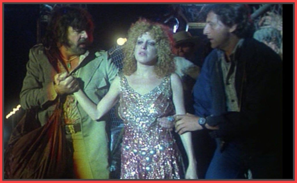 Woman in a glittery, sparkling gown is being escorted by two men who hold her arms in a dim backstage-like scene.