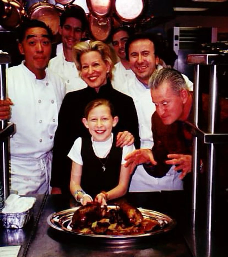 Smiling group of chefs and a woman posing with a young girl cutting a chocolate cake in a restaurant kitchen.