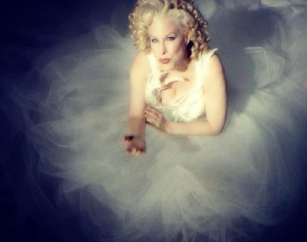 Woman in a white bridal gown with curly blonde hair, surrounded by wispy tulle or foggy fabric.