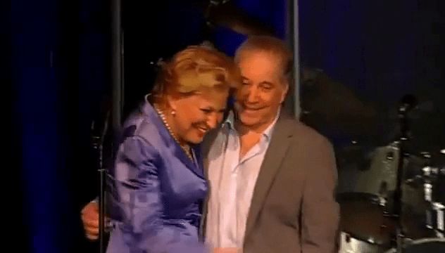 Bette Midler & Paul Simon smiling and embracing on a stage, the woman in a lavender dress and the man in a light jacket and shirt.