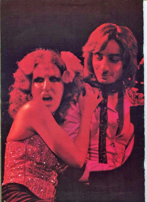 Fa40f60a7889025cf56ce31621364378 - bootleg betty Two performers on stage: a woman with curly blonde hair in a sparkling dress stretches toward the audience while a man in a light shirt stands beside her.