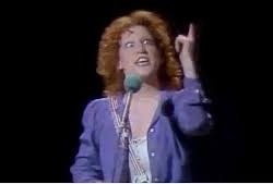 Female singer with curly red hair in a blue/purple blazer performing on stage, microphone in hand, pointing upward.