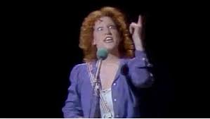 Female singer with curly red hair in a blue/purple blazer performing on stage, microphone in hand, pointing upward.