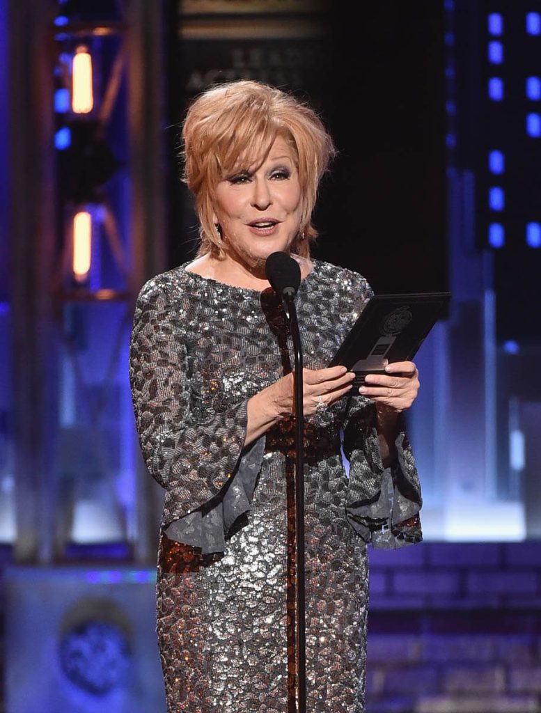 Older woman with short blonde hair in a sparkling silver gown speaks into a microphone on a stage, holding a booklet or card nearby.