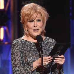 Older woman with short blonde hair in a sparkling silver gown speaks into a microphone on a stage, holding a booklet or card nearby.