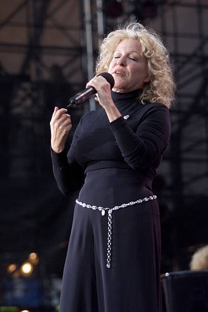 Female singer with curly blonde hair wearing a black outfit and silver belt performs on stage, holding a microphone.