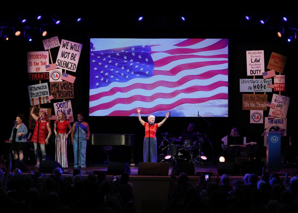 Stage performance with a? audience; singers at microphones, vibrant flag backdrop on screen, and protest signs around the sides.