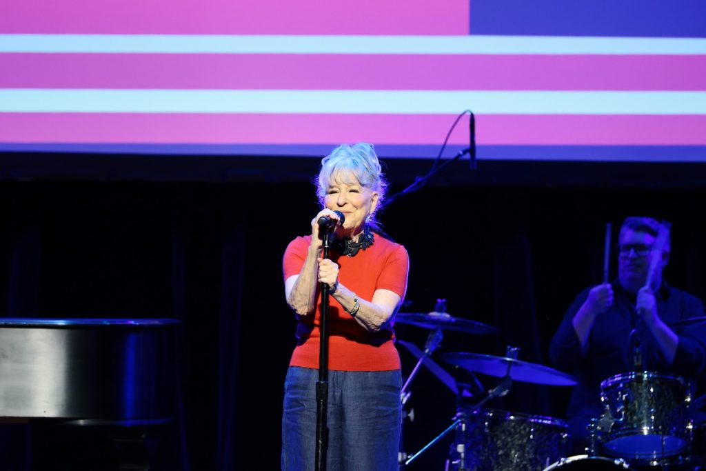 Older woman in a red top sings into a microphone on stage, with a drummer and purple-pink backdrop behind her.