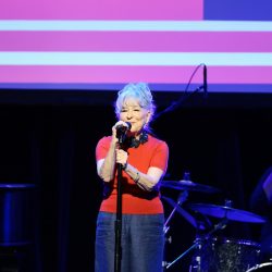 Older woman in a red top sings into a microphone on stage, with a drummer and purple-pink backdrop behind her.