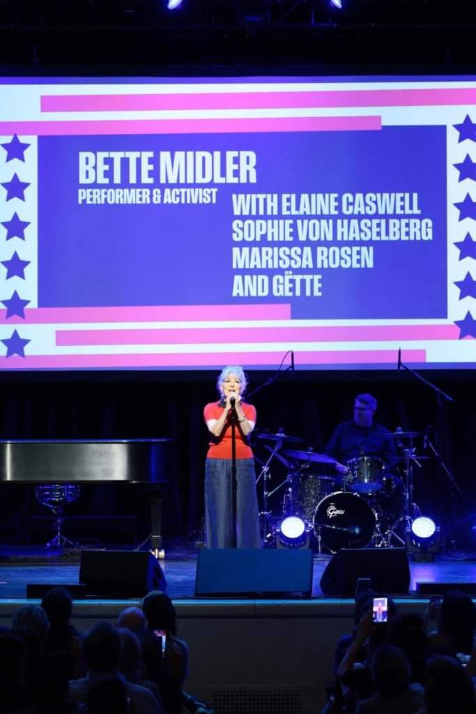 Bette Midler performing on stage with a large screen behind listing performers, including Elaine Caswell and Sophie von Haselberg, in blue and pink.