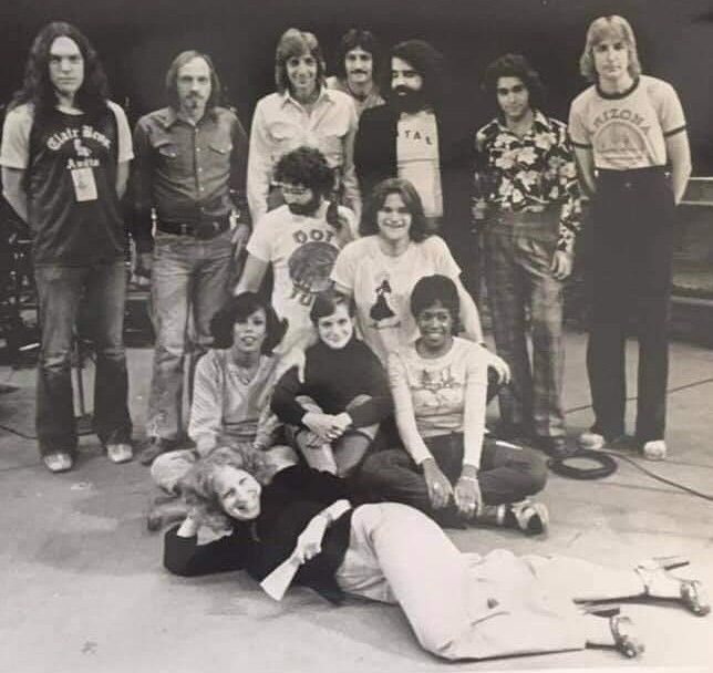 Black-and-white group photo of several people in 1970s-style clothing posing on a concrete surface; some stand, some sit or lie on the ground in front.