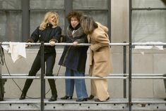 Three women leaning on a railing and looking down at something below, smiling as they chat.