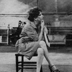 Black-and-white photo of a woman in a dress sitting on a small chair, elbows on knees, chin resting on her hands in an industrial-looking space