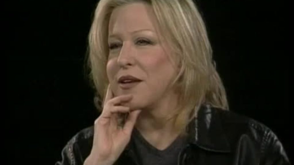 Woman with blonde hair rests her chin on her hand during a conversation, against a black background in a studio setup.