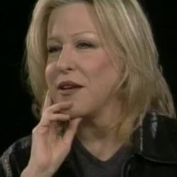 Woman with blonde hair rests her chin on her hand during a conversation, against a black background in a studio setup.