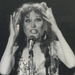 Female singer performing on stage, hands raised to her head, singing into a microphone in a floral dress (black-and-white photo).