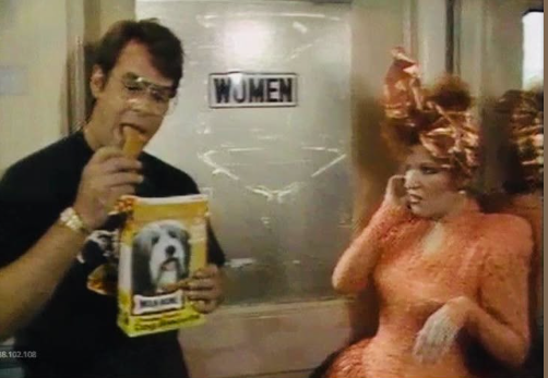 2026-06-19_11-32-18 - bootleg betty Two people in a backstage hallway near a women’s restroom door; a man is eating a snack from a box featuring a dog, while a woman in a pink dress with a feathered hat looks on.