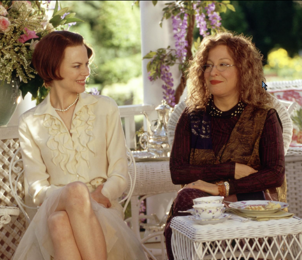Two women sit on a white wicker patio, chatting over tea amid flowers and greenery.