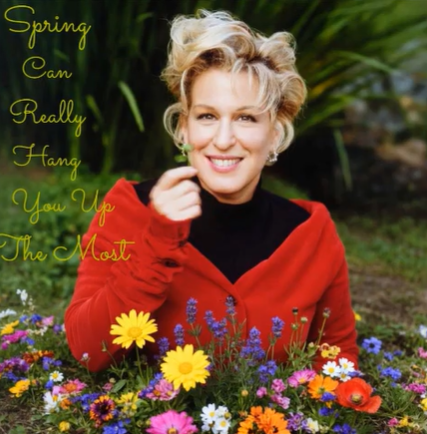 Smiling woman in a red cardigan leaning over a bed of colorful flowers, with green script text that reads 'Spring Can Really Hang You Up The Most' on the left.