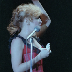 2026-07-08_11-31-06 - bootleg betty Female singer on stage, raising her right arm while performing into a microphone, wearing a red corset-style top.
