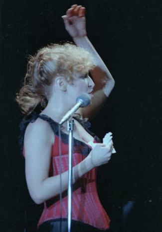 Female singer on stage, raising her right arm while performing into a microphone, wearing a red corset-style top.