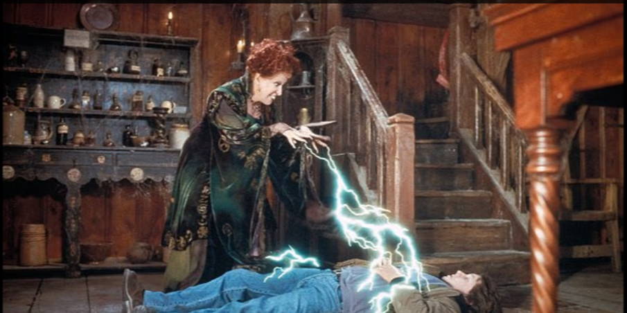 A witch in a green robe casts blue lightning at a man lying on the floor in a rustic wooden room.