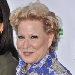 Older woman with short blond hair wearing a blue paisley-patterned shirt posing for a photo at an event, with a subtle smile and jewelry visible.