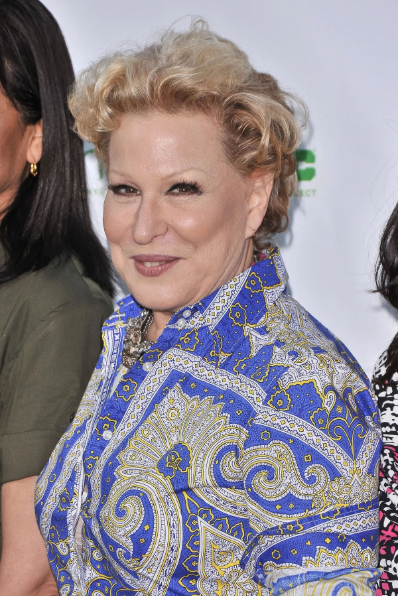 Older woman with short blond hair wearing a blue paisley-patterned shirt posing for a photo at an event, with a subtle smile and jewelry visible.