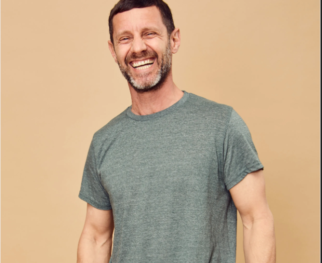 Smiling man in a gray tee stands against a beige background.