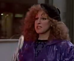 Bette Midler look?alike with curly red hair in a purple shiny windbreaker and black beret, carrying a light blue folder while walking on a city sidewalk.