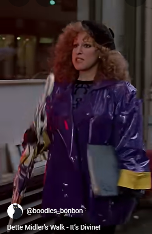 2026-07-19_13-00-10 - bootleg betty Bette Midler look?alike with curly red hair in a purple shiny windbreaker and black beret, carrying a light blue folder while walking on a city sidewalk.