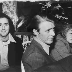 Three people sit at a table indoors: left man smiles at the camera, middle man looks to the side, woman on the right rests her chin on her hand.