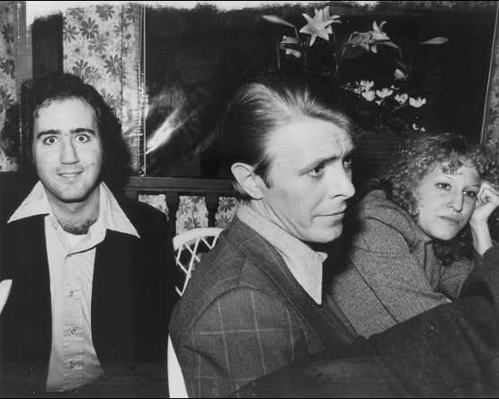 2026-07-20_06-27-30 - bootleg betty Three people sit at a table indoors: left man smiles at the camera, middle man looks to the side, woman on the right rests her chin on her hand.