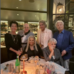 Six adults posing for a group photo at a restaurant table with wine glasses and warm lighting in the background.
