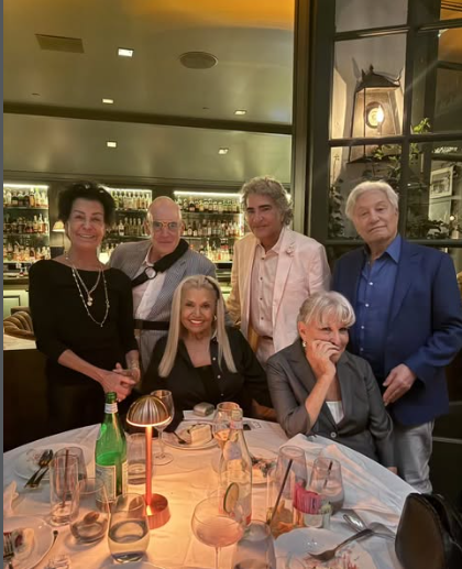 Six adults posing for a group photo at a restaurant table with wine glasses and warm lighting in the background.