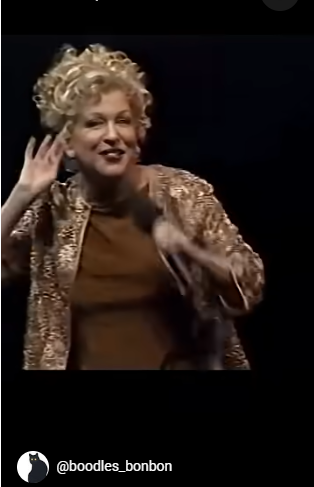 2026-07-26_14-04-27 - bootleg betty Female performer with short curly blonde hair in a gold sequin jacket, smiling and cupping her ear while performing on a dark stage.