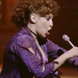 Female singer in a torn purple top performs into a microphone on stage (side profile).