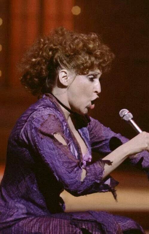 Female singer in a torn purple top performs into a microphone on stage (side profile).