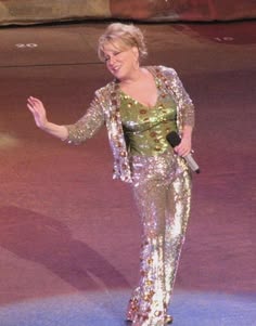Female singer on stage in a sparkling sequined suit, waving with a microphone in hand under stage lights.
