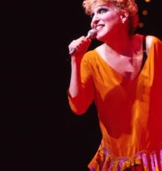 Female singer in an orange dress performing on stage, holding a microphone under warm stage lights.