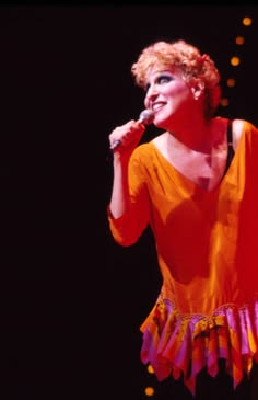 Female singer in an orange dress performing on stage, holding a microphone under warm stage lights.