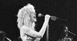 Black-and-white photo of a female vocalist with curly hair singing into a microphone on stage, with a guitarist visible in the background.