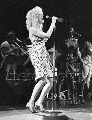 Black-and-white photo of a female vocalist with curly hair singing into a microphone on stage, with a guitarist visible in the background.
