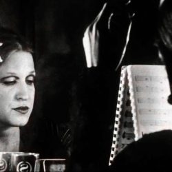 Black-and-white image of a woman smoking a cigarette, gazing at her reflection in a mirror with a music stand and sheet music nearby.