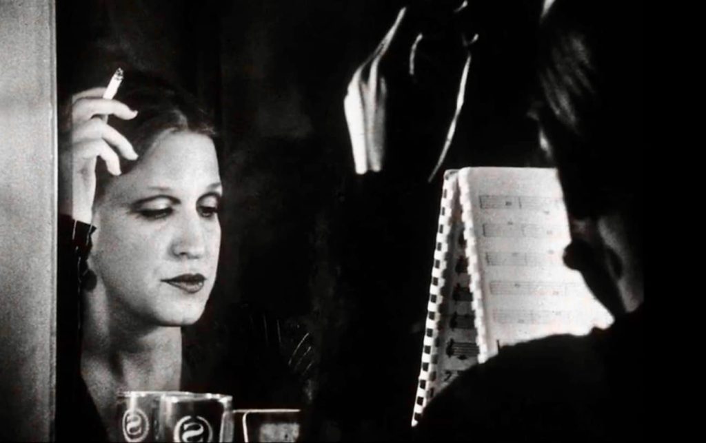 Black-and-white image of a woman smoking a cigarette, gazing at her reflection in a mirror with a music stand and sheet music nearby.
