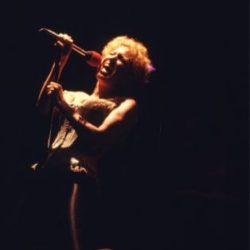 43d0b97476d05a116d2b6f407d579f44 - bootleg betty Female singer kneels on stage, singing into a microphone under dramatic spotlight.